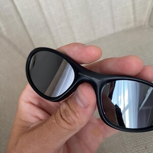 Puede incluir: Gafas de sol negras con lentes reflectantes. Las gafas tienen un dise&ntilde;o elegante y curvado, y el nombre de la marca "Oakley" es visible en la montura. Las lentes son oscuras y reflejan el entorno.