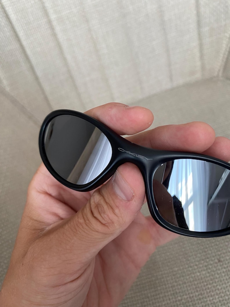 Puede incluir: Gafas de sol negras con lentes reflectantes. Las gafas tienen un dise&ntilde;o elegante y curvado, y el nombre de la marca "Oakley" es visible en la montura. Las lentes son oscuras y reflejan el entorno.