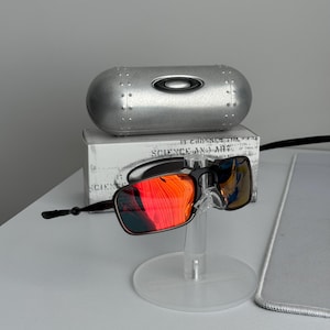 May include: Black and red sunglasses with a silver case. The sunglasses have a rectangular frame and red-tinted lenses. The case is metallic and oval-shaped. The sunglasses are displayed on a clear stand.