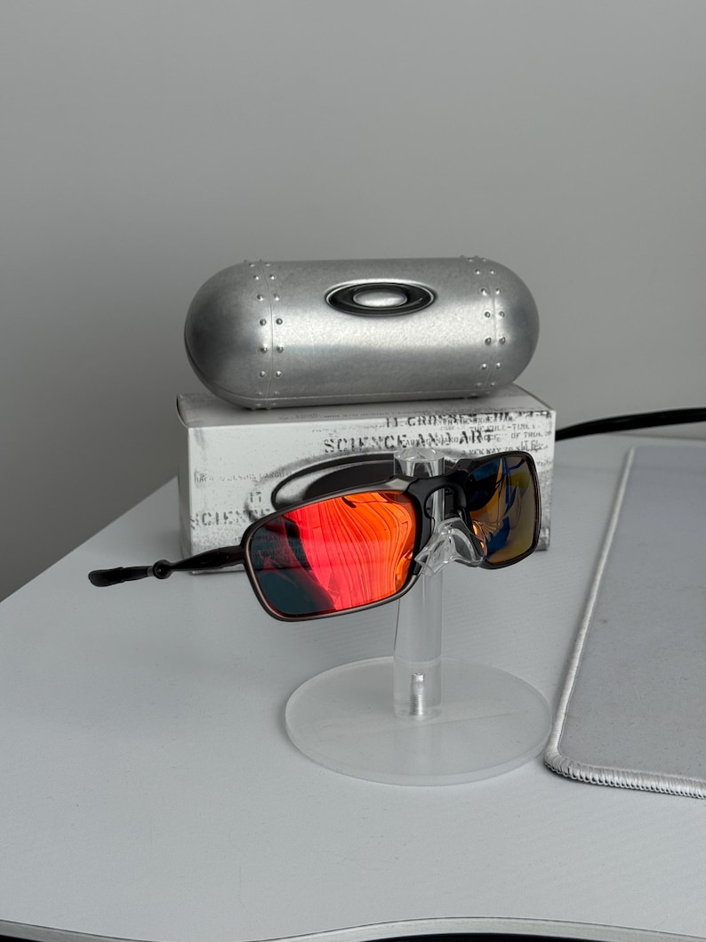 May include: Black and red sunglasses with a silver case. The sunglasses have a rectangular frame and red-tinted lenses. The case is metallic and oval-shaped. The sunglasses are displayed on a clear stand.
