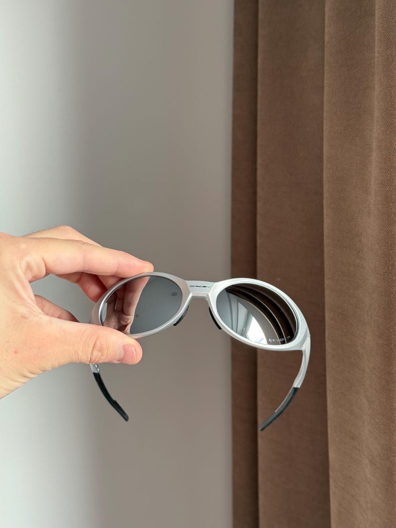 May include: Silver sunglasses with dark lenses. The sunglasses have a modern design with curved arms and black earpieces. The lenses reflect the surrounding environment.
