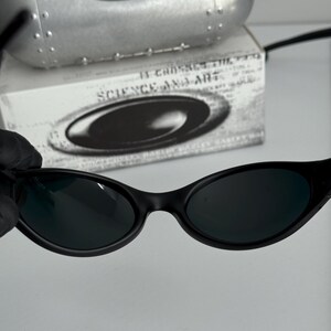 May include: Black oval-shaped sunglasses with dark lenses. The sunglasses are set against a white background, with a silver case and a box with the text "SCIENCE AND ART". The sunglasses have a sleek, modern design.
