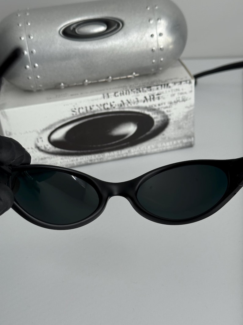 May include: Black oval-shaped sunglasses with dark lenses. The sunglasses are set against a white background, with a silver case and a box with the text "SCIENCE AND ART". The sunglasses have a sleek, modern design.