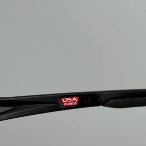 Puede incluir: Primer plano de una montura de gafas negra con el texto "USA ASSEMBLED" en un rect&aacute;ngulo rojo. La montura est&aacute; sobre un fondo gris. La montura tambi&eacute;n tiene el texto "CE" y "UK".