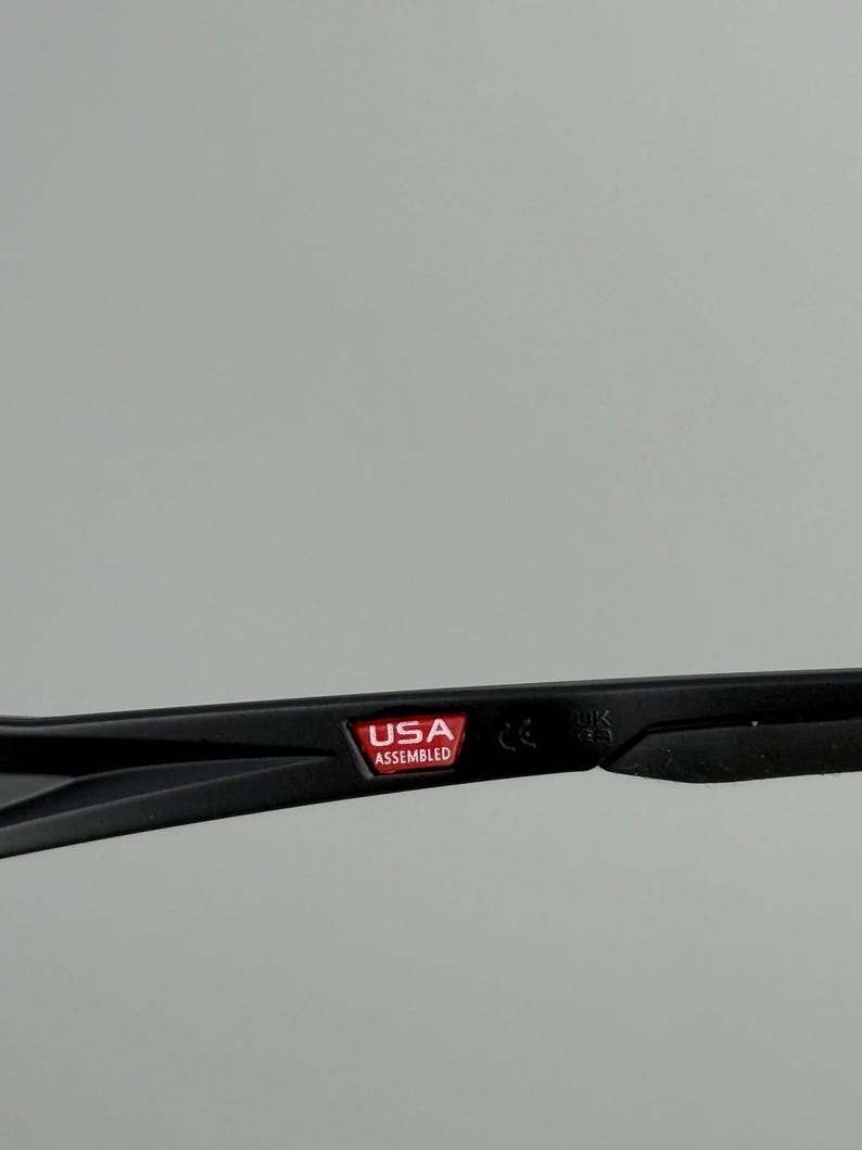 Puede incluir: Primer plano de una montura de gafas negra con el texto "USA ASSEMBLED" en un rect&aacute;ngulo rojo. La montura est&aacute; sobre un fondo gris. La montura tambi&eacute;n tiene el texto "CE" y "UK".