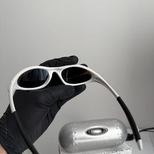 May include: White framed sunglasses with dark lenses. The sunglasses are held by a gloved hand, with a silver case and a box with the text "SCIENCE AND ART" visible in the background. The sunglasses have a sleek, modern design.