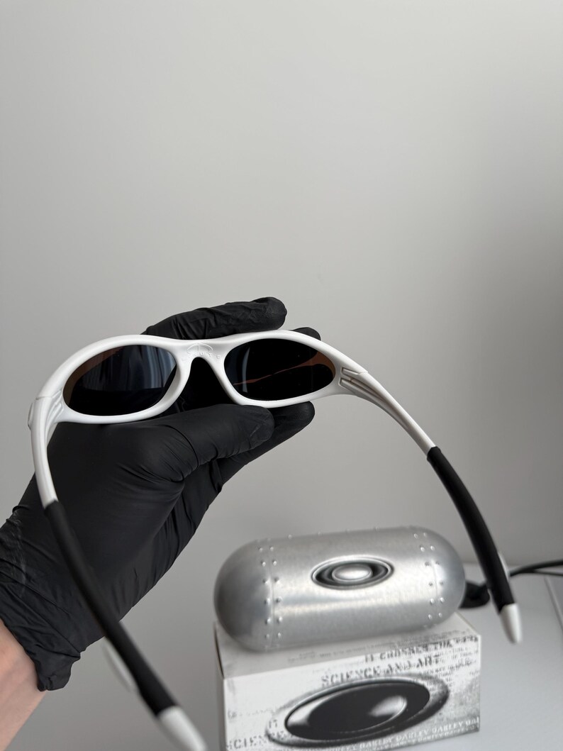 May include: White framed sunglasses with dark lenses. The sunglasses are held by a gloved hand, with a silver case and a box with the text "SCIENCE AND ART" visible in the background. The sunglasses have a sleek, modern design.