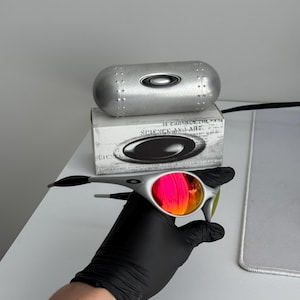 Pu&ograve; includere: Occhiali da sole argentati con lenti rosa e arancioni. Gli occhiali sono tenuti vicino a un astuccio argentato e a una scatola con la scritta "SCIENCE AND ART". Le lenti riflettono la luce e le aste sono nere.