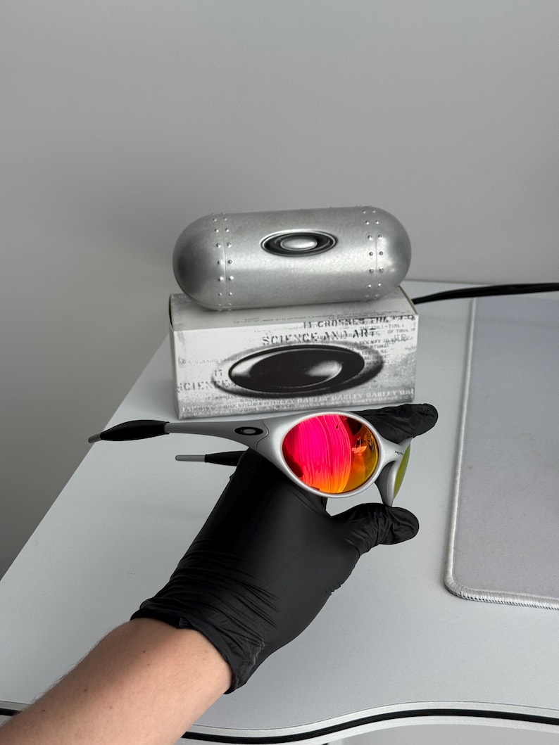 Pu&ograve; includere: Occhiali da sole argentati con lenti rosa e arancioni. Gli occhiali sono tenuti vicino a un astuccio argentato e a una scatola con la scritta "SCIENCE AND ART". Le lenti riflettono la luce e le aste sono nere.