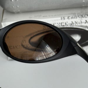 Puede incluir: Gafas de sol negras con lentes marrones. Las gafas tienen un dise&ntilde;o elegante y curvado. Las lentes tienen un ligero tinte y reflejan la luz. La montura es negra y parece estar hecha de un material duradero. El texto "PRIZM" es visible en la lente.