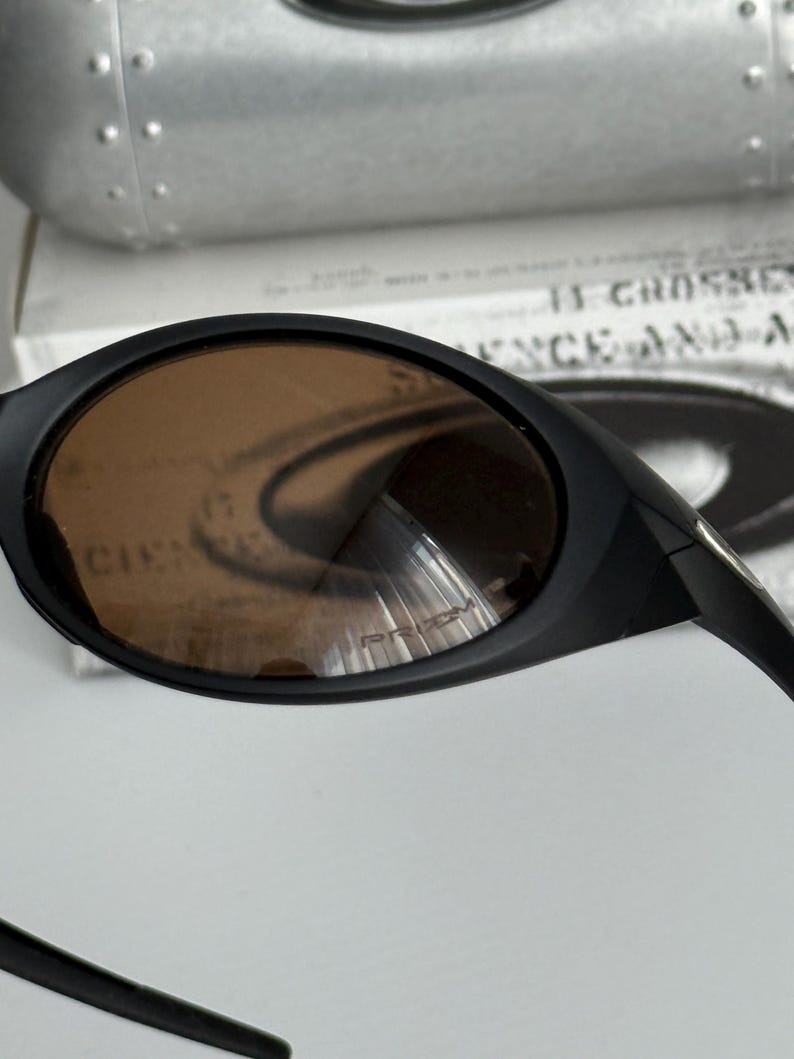 Puede incluir: Gafas de sol negras con lentes marrones. Las gafas tienen un dise&ntilde;o elegante y curvado. Las lentes tienen un ligero tinte y reflejan la luz. La montura es negra y parece estar hecha de un material duradero. El texto "PRIZM" es visible en la lente.
