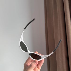 以下が含まれることがあります： 白いフレームと濃い色のレンズのサングラス。 サングラスは、テンプルに黒いアクセントが付いた、洗練されたモダンなデザインです。 フレームは軽量素材で作られているようです。
