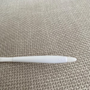 Puede incluir: Un test de embarazo de pl&aacute;stico blanco con una punta redondeada y una serie de tres peque&ntilde;as flechas. El test est&aacute; sobre una superficie texturizada de color gris claro.