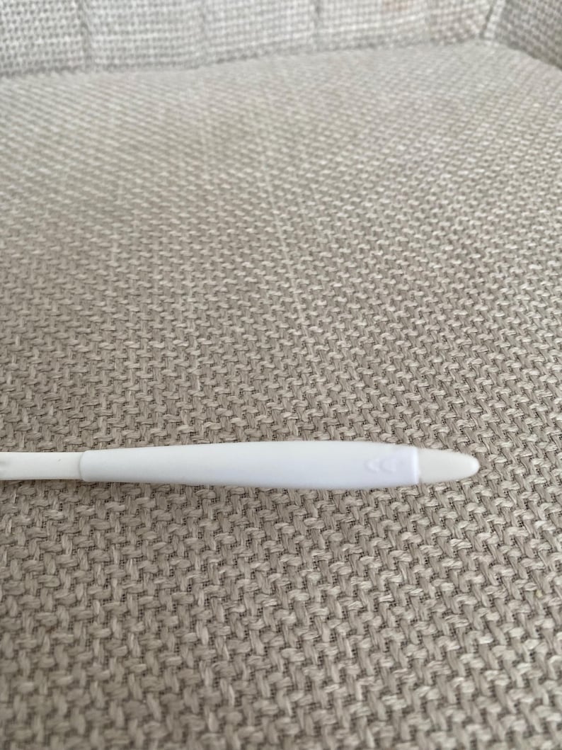 Puede incluir: Un test de embarazo de pl&aacute;stico blanco con una punta redondeada y una serie de tres peque&ntilde;as flechas. El test est&aacute; sobre una superficie texturizada de color gris claro.