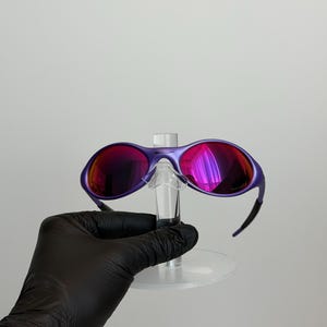 May include: Purple sunglasses with red-tinted lenses. The sunglasses have a sleek, modern design with a curved frame and black arms. The lenses reflect a vibrant, multi-colored sheen. The sunglasses are displayed on a clear stand.