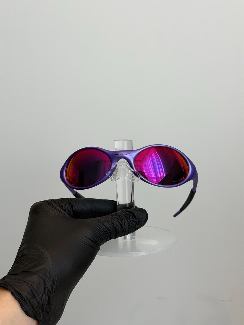 May include: Purple sunglasses with red-tinted lenses. The sunglasses have a sleek, modern design with a curved frame and black arms. The lenses reflect a vibrant, multi-colored sheen. The sunglasses are displayed on a clear stand.