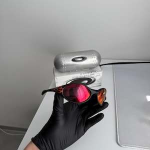 May include: Black sunglasses with red mirrored lenses and a flame design on the temples. The sunglasses are held in a black gloved hand. A silver case and box are in the background.