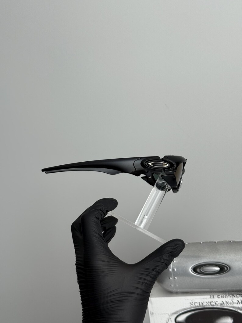 May include: Black sunglasses with a sleek, modern design. The sunglasses are held up by a clear stand. The lenses are dark, and the frame is a glossy black. The overall aesthetic is sporty and stylish.