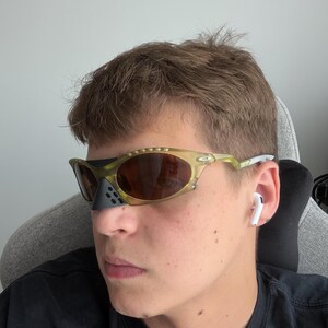 May include: Olive green and brown sunglasses with a black nose guard. The sunglasses have a unique, sporty design. The lenses are brown and reflective. The frame is a mix of olive green and clear plastic.