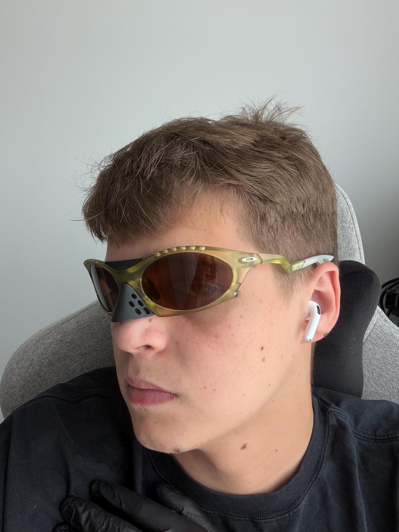May include: Olive green and brown sunglasses with a black nose guard. The sunglasses have a unique, sporty design. The lenses are brown and reflective. The frame is a mix of olive green and clear plastic.