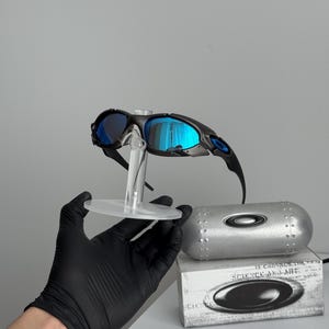 May include: Black framed sunglasses with blue mirrored lenses are displayed on a clear stand. A silver case and a box with the text "SCIENCE AND ART" are also visible.