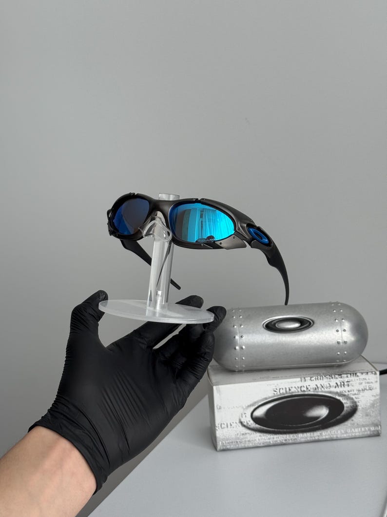 May include: Black framed sunglasses with blue mirrored lenses are displayed on a clear stand. A silver case and a box with the text "SCIENCE AND ART" are also visible.