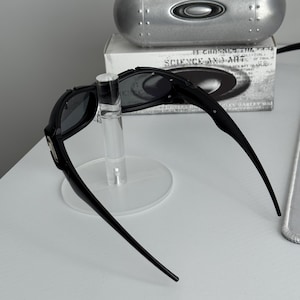 May include: Black sunglasses with a sleek design, displayed on a clear stand. The sunglasses have dark lenses and a curved frame. A silver case and a box with text are in the background.