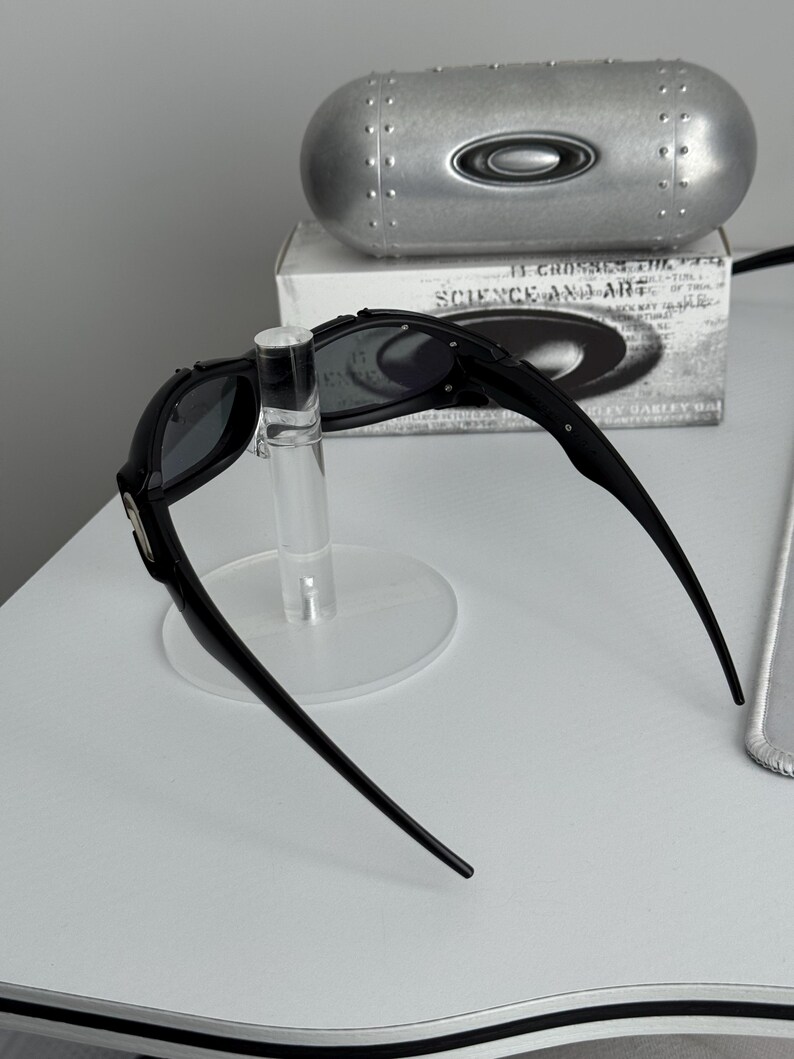 May include: Black sunglasses with a sleek design, displayed on a clear stand. The sunglasses have dark lenses and a curved frame. A silver case and a box with text are in the background.