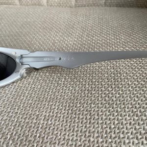 May include: Gray sunglasses with dark lenses. The frame is a matte gray color, and the temples are slender and curved. The text "MADE IN U.S.A." is printed on the temple. The sunglasses are resting on a textured, neutral-colored surface.