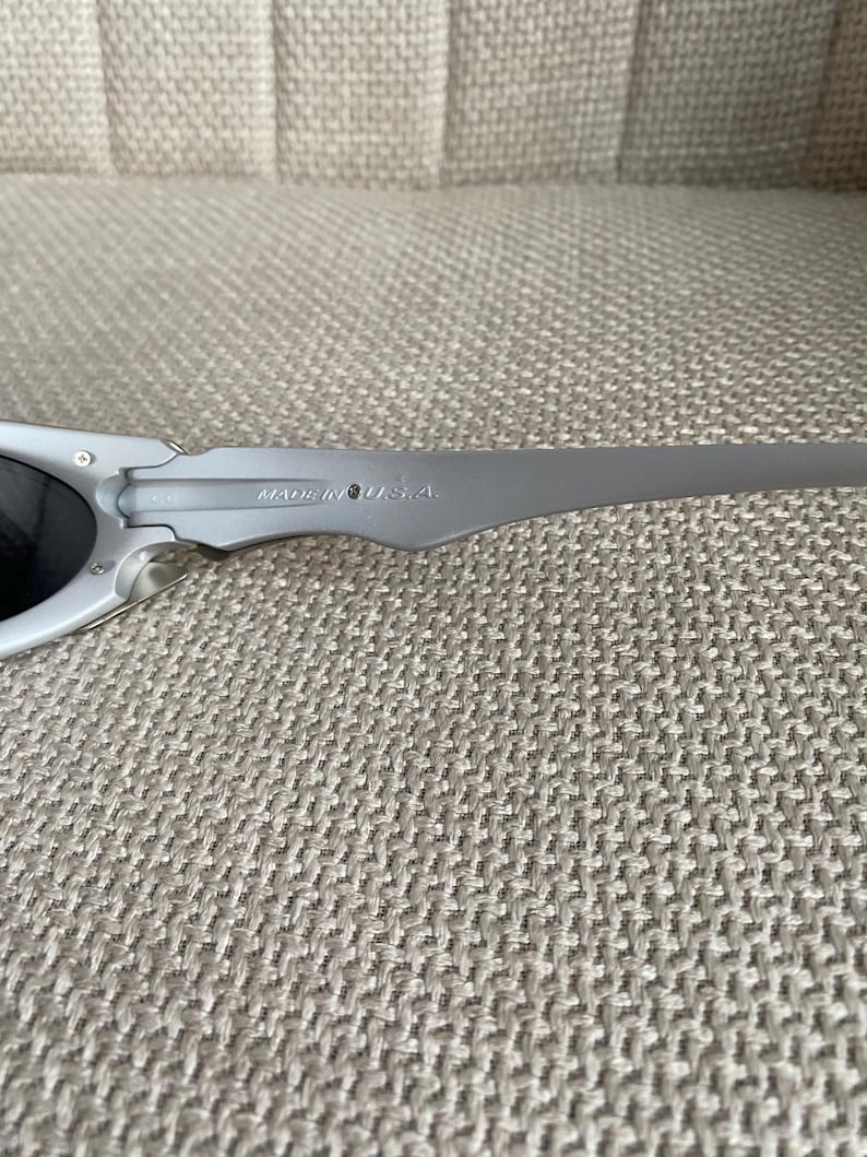 May include: Gray sunglasses with dark lenses. The frame is a matte gray color, and the temples are slender and curved. The text "MADE IN U.S.A." is printed on the temple. The sunglasses are resting on a textured, neutral-colored surface.