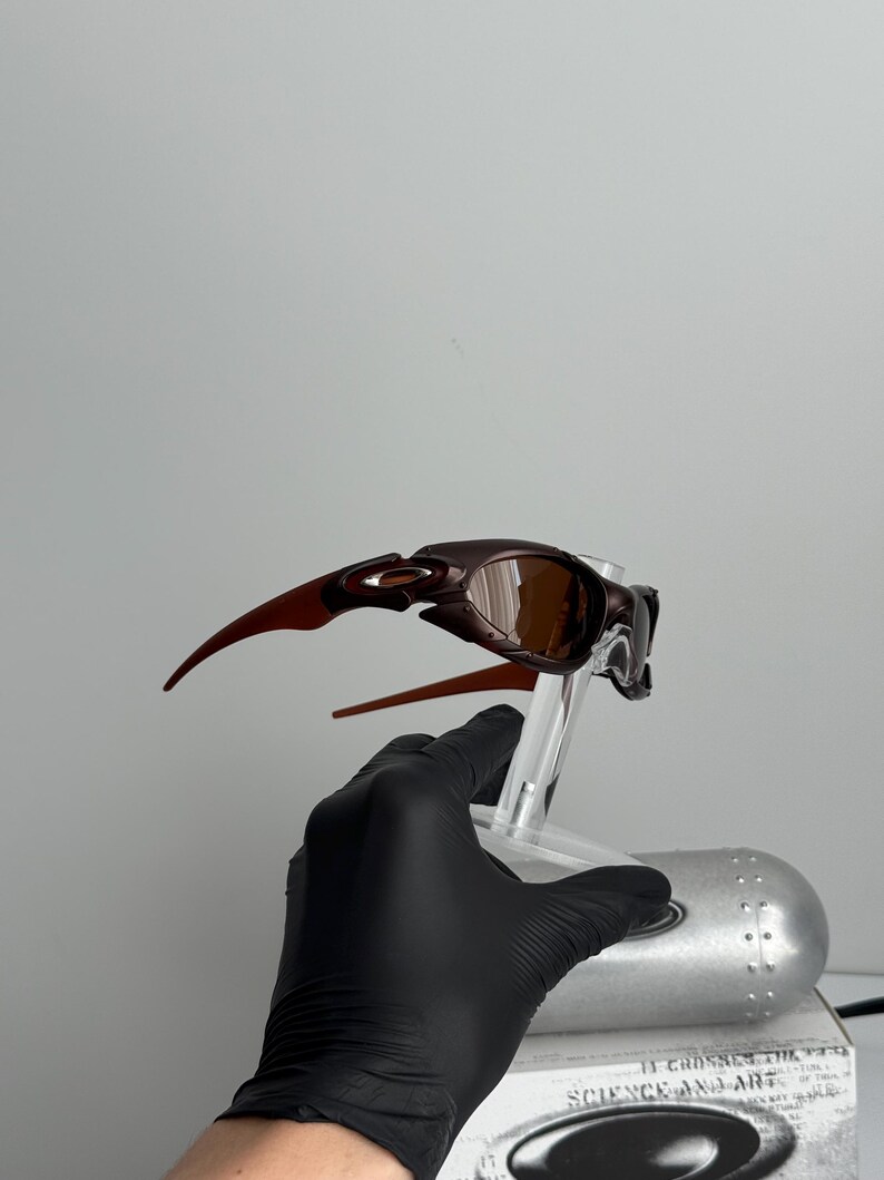 May include: Brown and bronze-colored sunglasses with a sleek, modern design. The sunglasses have a curved frame and lenses, and are held by a clear stand. The image is set against a plain background.