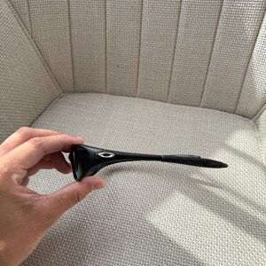 May include: Black sunglasses with a streamlined design. The sunglasses feature a curved frame and dark lenses. The Oakley logo is visible on the temples. The sunglasses are held in a person's hand.