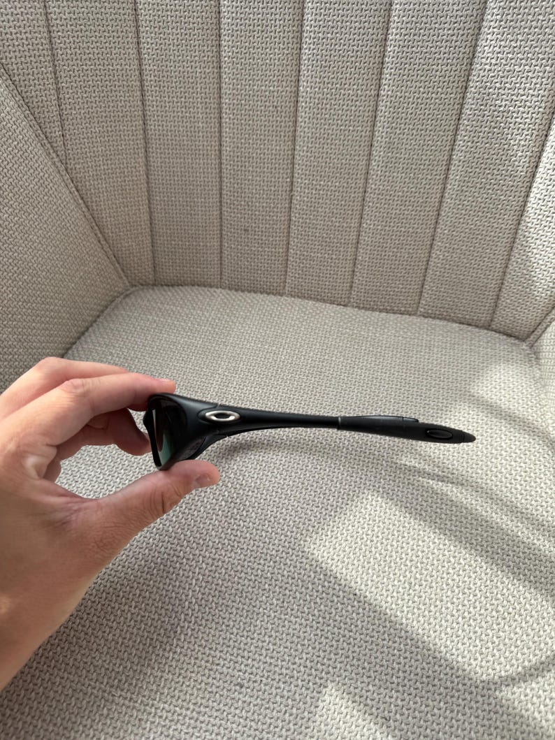 May include: Black sunglasses with a streamlined design. The sunglasses feature a curved frame and dark lenses. The Oakley logo is visible on the temples. The sunglasses are held in a person's hand.