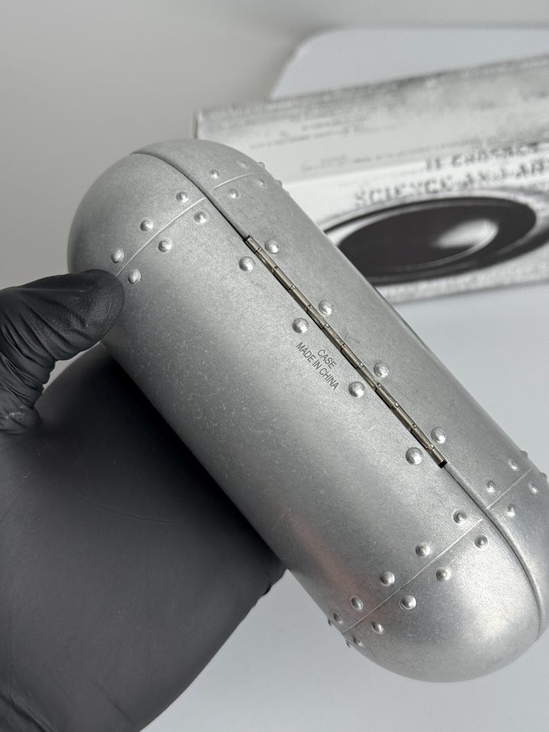 May include: A silver metal case with a riveted design and a rounded rectangular form. The case is hinged and features the text "MADE IN CHINA". A box in the background displays the words "SCIENCE AND ART".