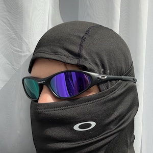 May include: Black Oakley sunglasses with purple and green lenses are worn over a black balaclava. The balaclava has a white Oakley logo on the front. The sunglasses have a sleek, sporty design.