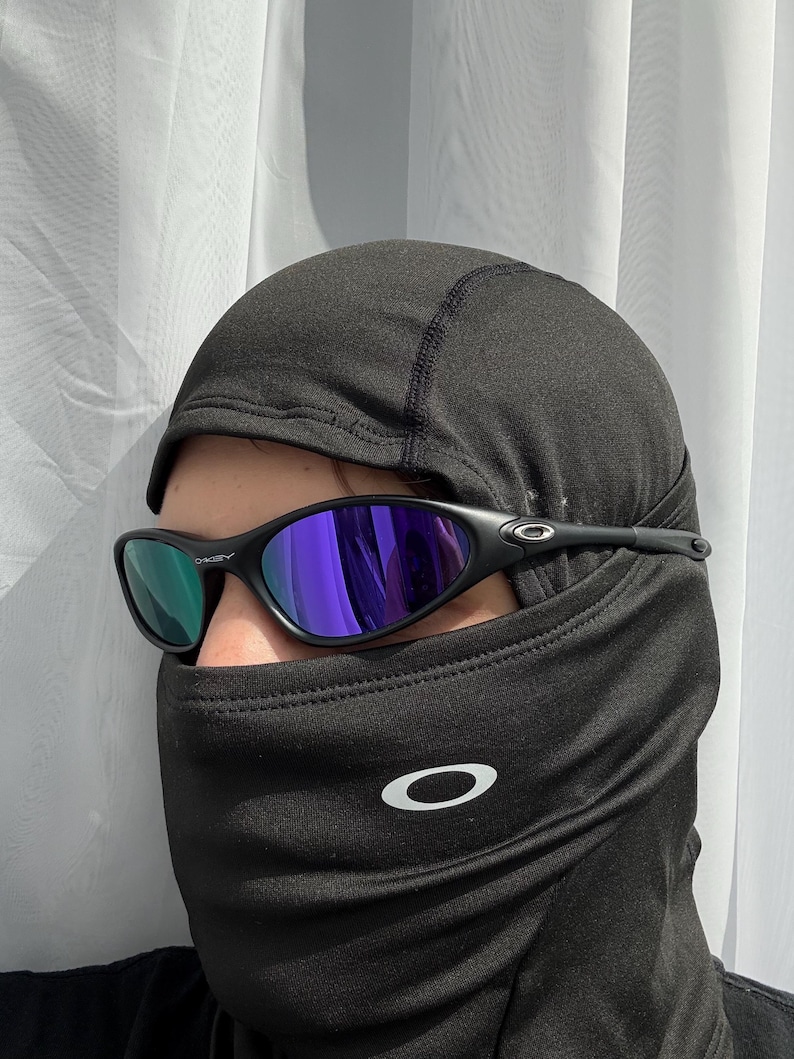 May include: Black Oakley sunglasses with purple and green lenses are worn over a black balaclava. The balaclava has a white Oakley logo on the front. The sunglasses have a sleek, sporty design.