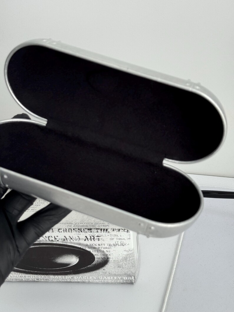 May include: Open silver eyeglasses case with a black interior. The case is oval-shaped and has a hinged design. The case is empty and ready to hold a pair of glasses. The case is sitting on top of a box with text.