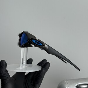 May include: Black and gray sunglasses with blue lenses. The sunglasses have a sleek, modern design with a curved frame and a small, circular logo on the arm. The lenses are a vibrant blue color.