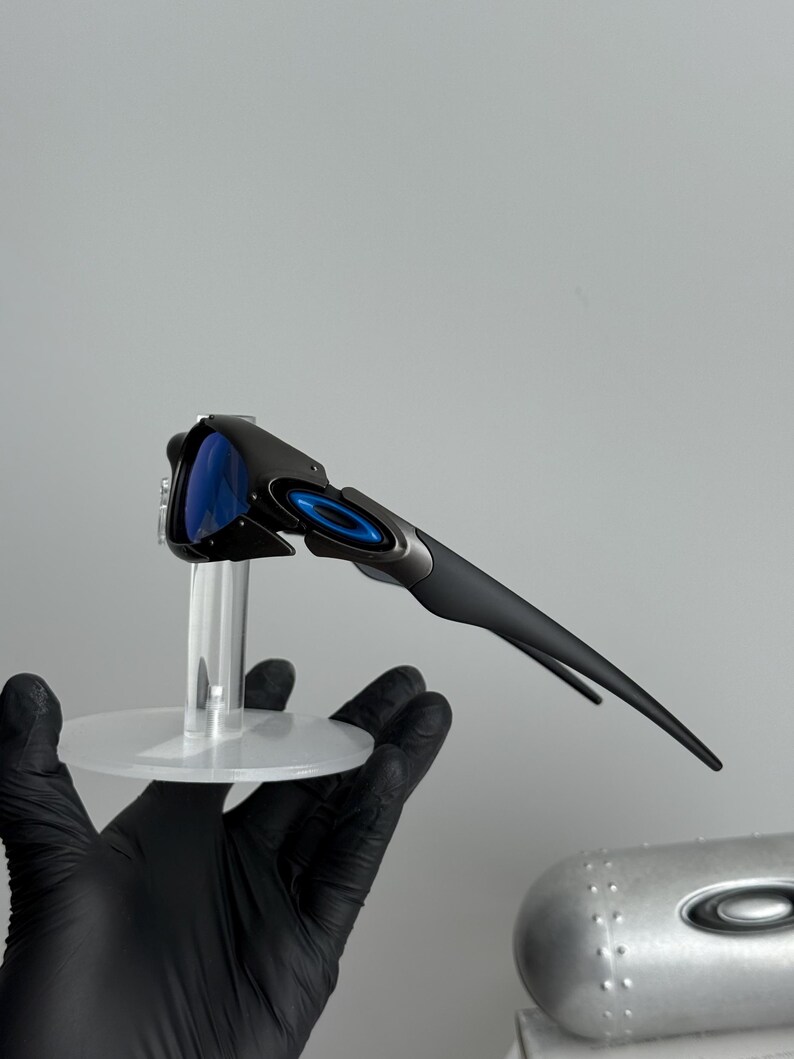 May include: Black and gray sunglasses with blue lenses. The sunglasses have a sleek, modern design with a curved frame and a small, circular logo on the arm. The lenses are a vibrant blue color.