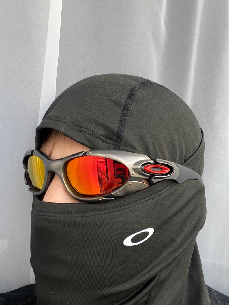 Op de afbeelding: Zilverkleurige en rood getinte zonnebril met een zwart montuur. De zonnebril heeft een sportief design met een rode accent op de poot. De lenzen reflecteren een gele en rode tint. De persoon draagt een zwarte bivakmuts.