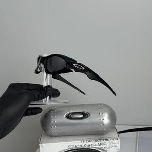May include: Black Oakley sunglasses with a unique, angular design, displayed on a clear stand. A silver case and a white box with the text "SCIENCE AND ART" are also present. The sunglasses have a sleek, modern aesthetic.