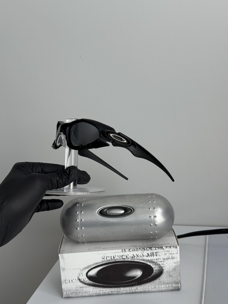 May include: Black Oakley sunglasses with a unique, angular design, displayed on a clear stand. A silver case and a white box with the text "SCIENCE AND ART" are also present. The sunglasses have a sleek, modern aesthetic.