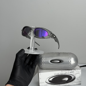 May include: Gray sunglasses with purple lenses are displayed on a clear stand. A silver case and a white box with the text "SCIENCE AND ART" are also present. The sunglasses have a sleek, modern design.