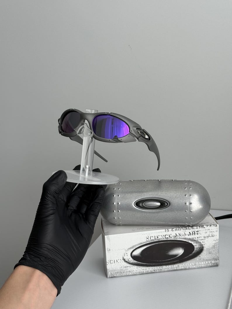 May include: Gray sunglasses with purple lenses are displayed on a clear stand. A silver case and a white box with the text "SCIENCE AND ART" are also present. The sunglasses have a sleek, modern design.