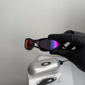May include: Black sunglasses with a sleek, curved frame and multi-colored lenses. The lenses shift from red to purple and blue. The sunglasses are held by a gloved hand, with a silver case and box in the background.