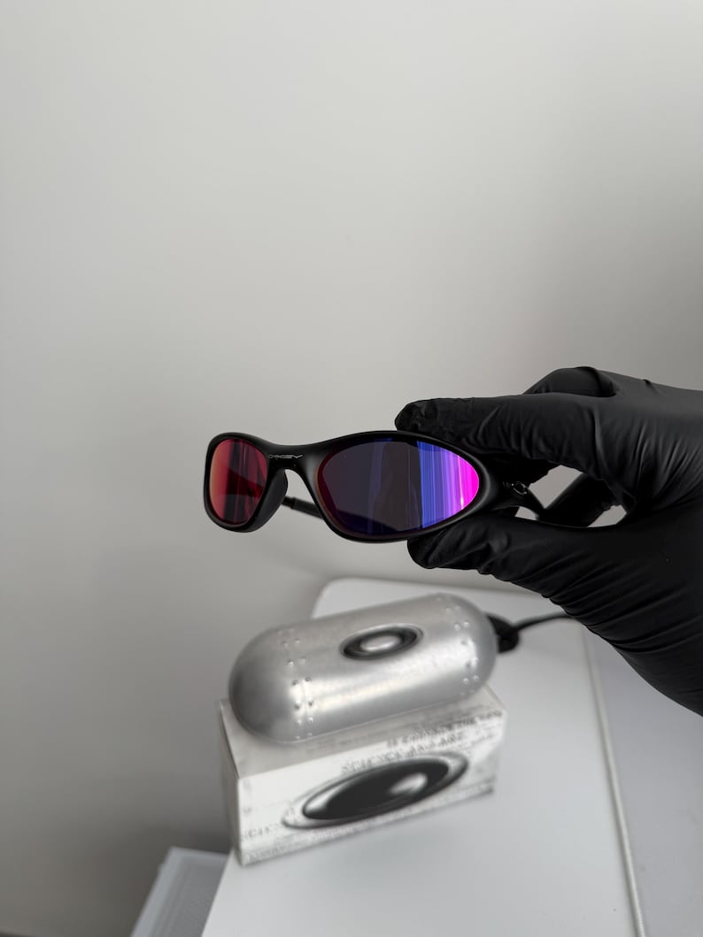 May include: Black sunglasses with a sleek, curved frame and multi-colored lenses. The lenses shift from red to purple and blue. The sunglasses are held by a gloved hand, with a silver case and box in the background.