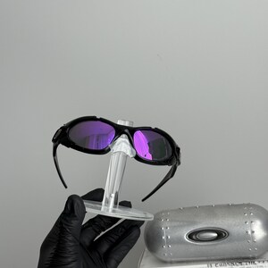 May include: Black sunglasses with purple lenses are displayed on a clear stand. The sunglasses have a sporty design. A silver case and a white box with text are also visible.