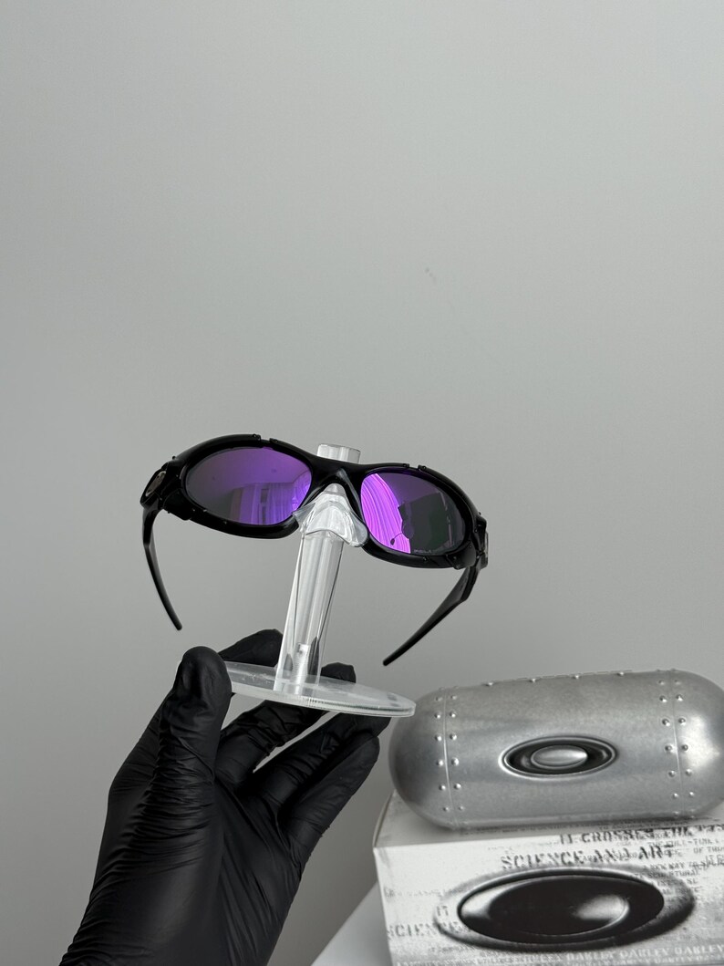 May include: Black sunglasses with purple lenses are displayed on a clear stand. The sunglasses have a sporty design. A silver case and a white box with text are also visible.