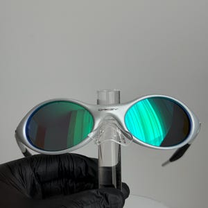 May include: Silver sunglasses with green mirrored lenses. The sunglasses have a modern design with a bridge and arms. The lenses reflect a vibrant green hue, and the frame has the brand name "Oakley" printed on the top.