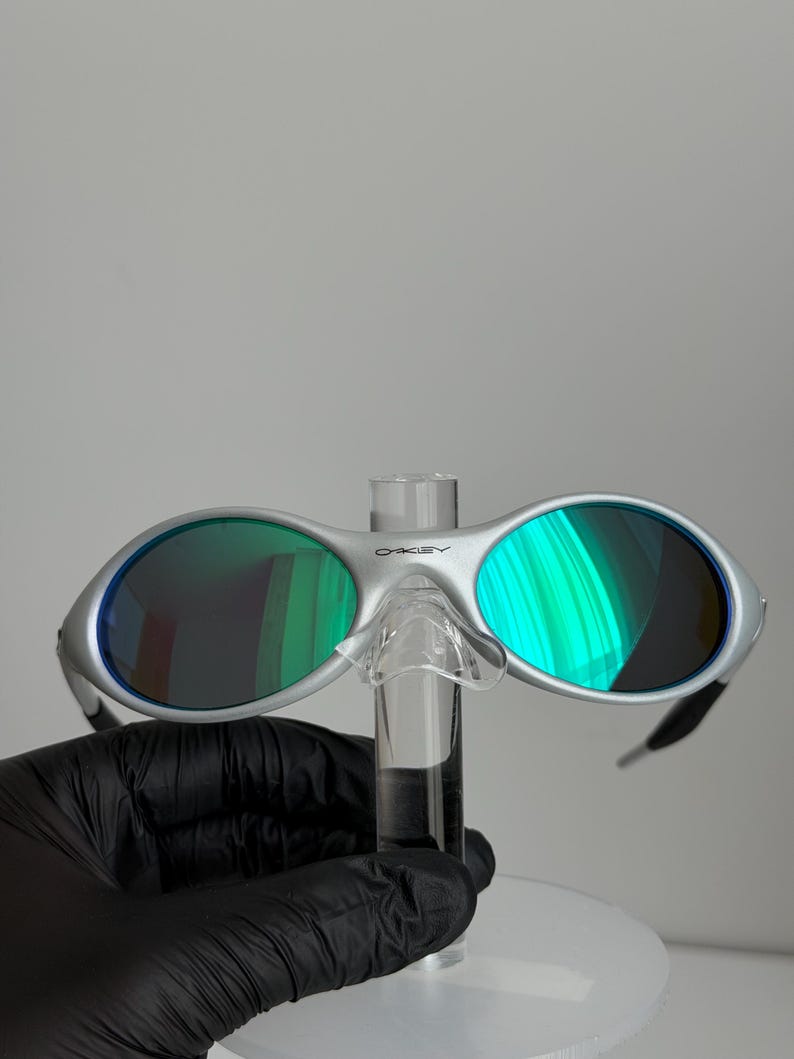 May include: Silver sunglasses with green mirrored lenses. The sunglasses have a modern design with a bridge and arms. The lenses reflect a vibrant green hue, and the frame has the brand name "Oakley" printed on the top.